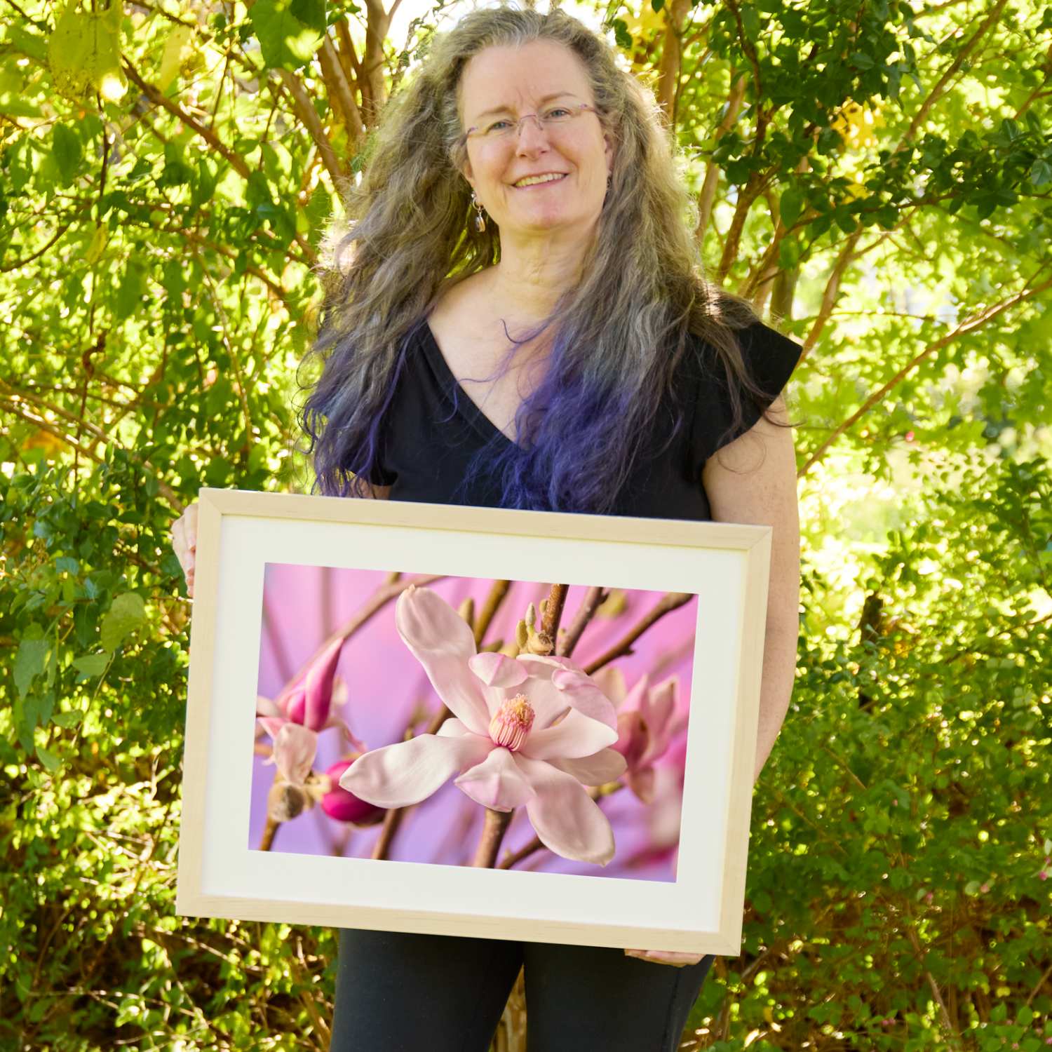 Christina Peters holding Magnolia Whisper Wall Art Print, a horizontal close-up of a pale pink magnolia blossom and magenta buds on slender branches against a soft pink backdrop, framed in natural wood with a white mat. She is standing outdoors in front of sunlit green trees and leafy shrubs.