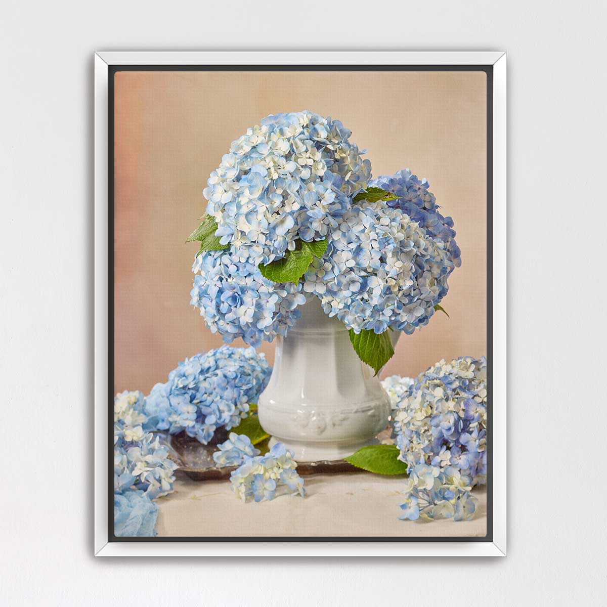 A white vase filled with blue hydrangea flowers sits on a table, surrounded by additional blue hydrangea flowers.