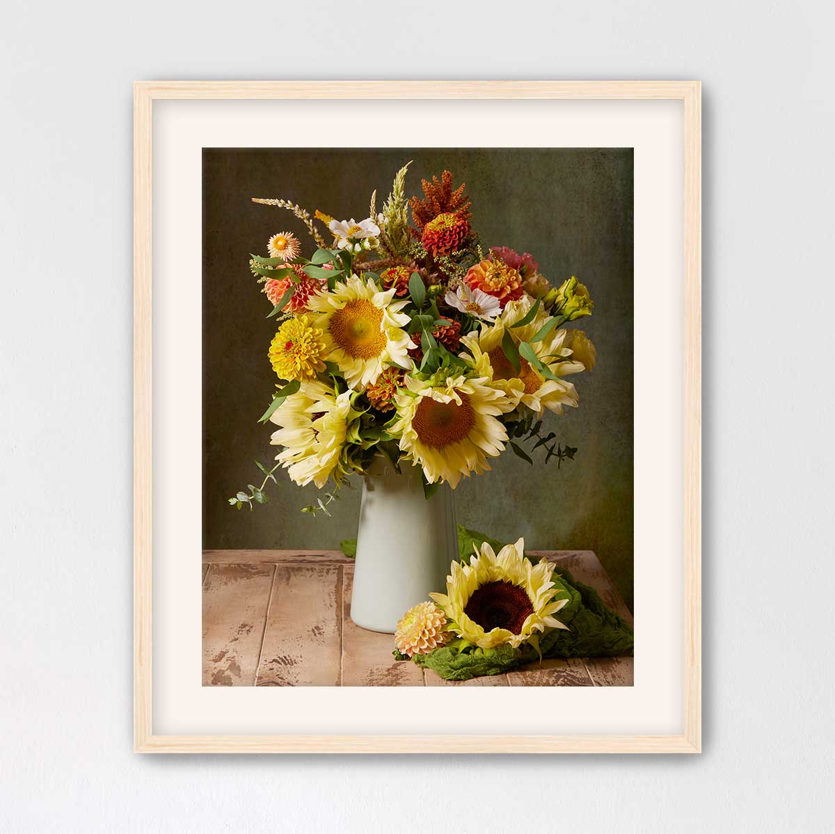 A framed still life photograph depicting a vase filled with a vibrant bouquet of yellow sunflowers and other flowers, placed on a wooden surface.