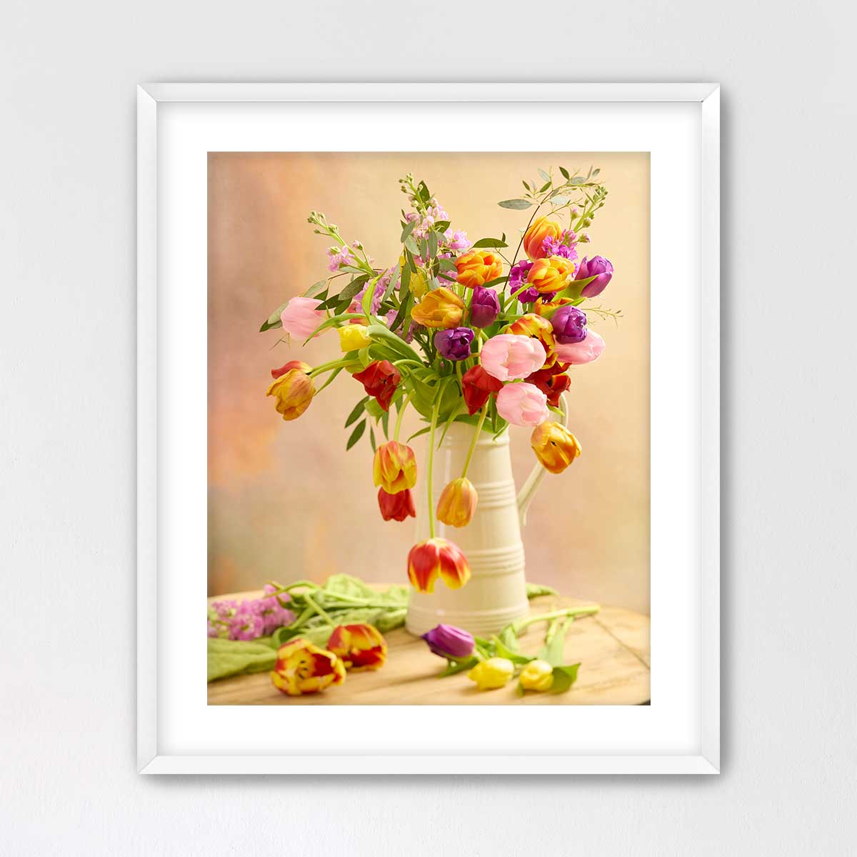 A white vase filled with a vibrant bouquet of tulips sits on a wooden surface, surrounded by additional flowers and foliage.