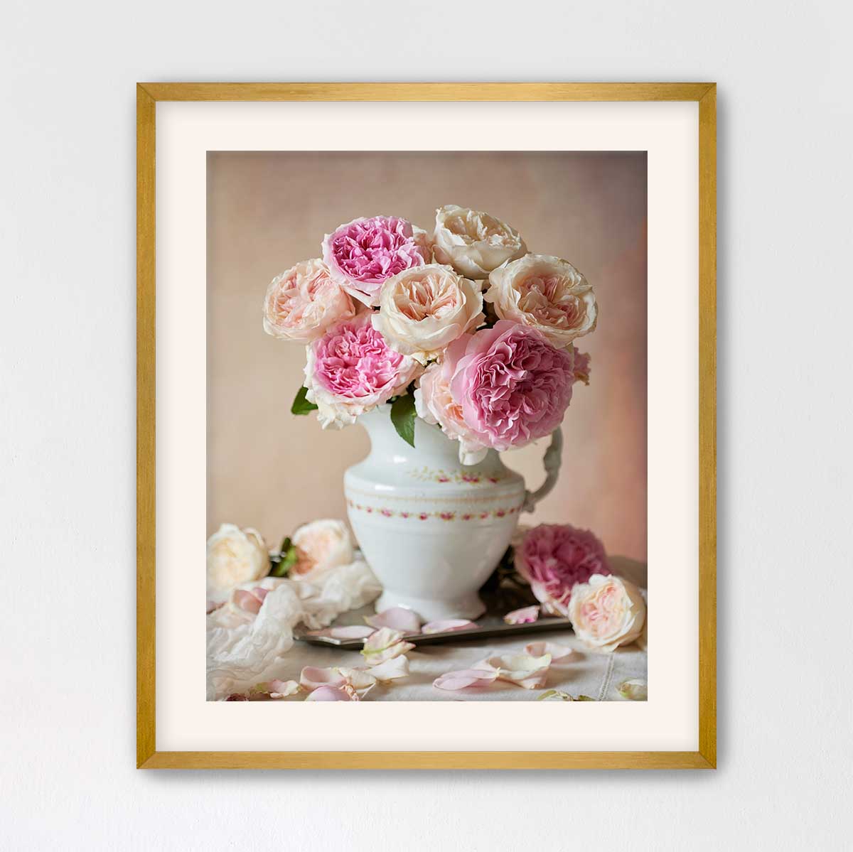 A white vase with pink and white roses sits on a table, surrounded by additional rose petals.