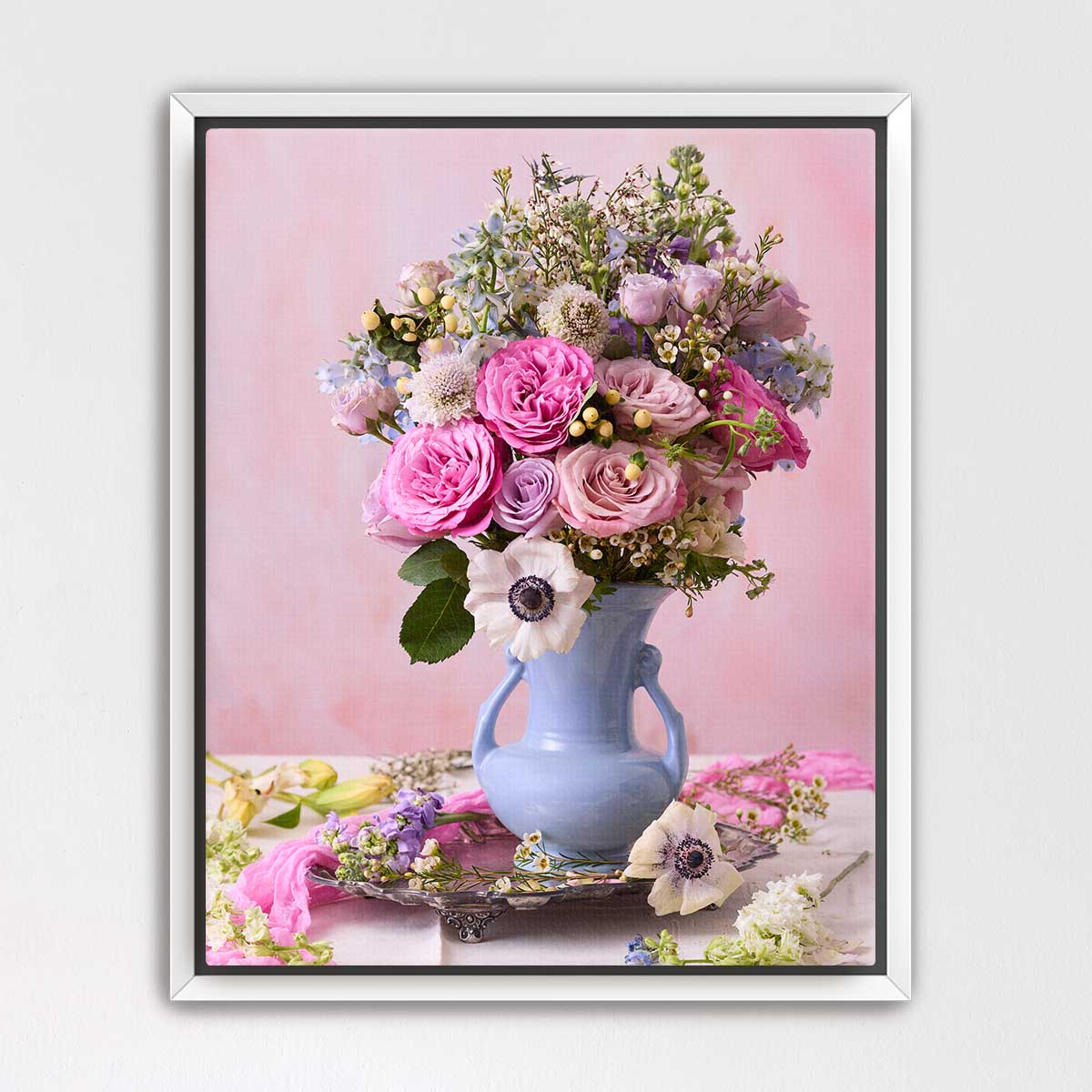 A bouquet of pink roses and white anemones is displayed in a blue vase on a pink surface, creating a visually appealing floral arrangement.