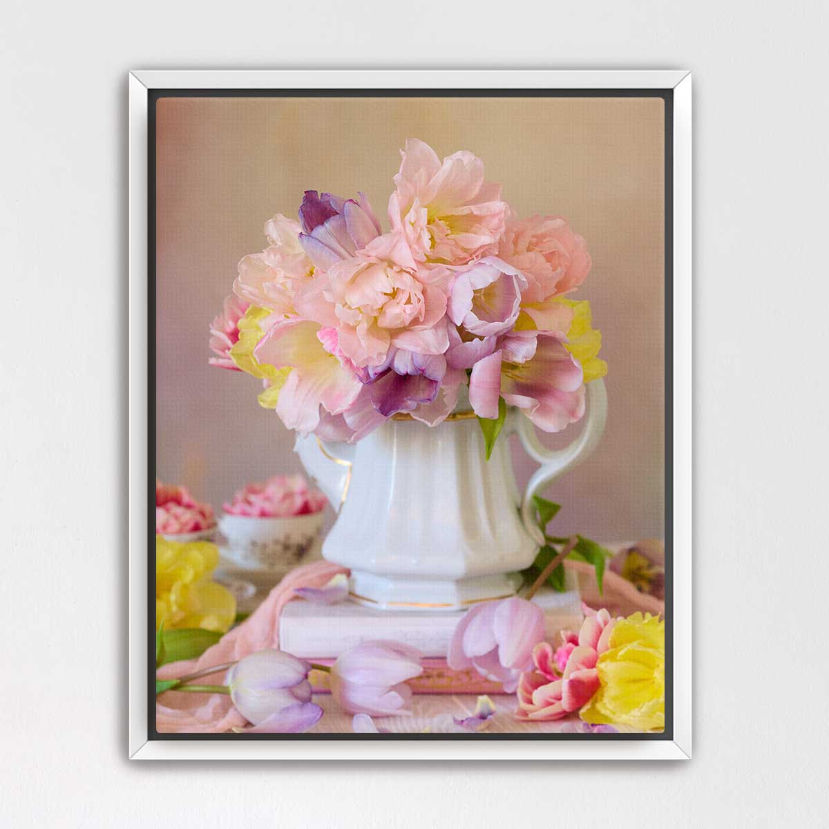 A white vase filled with a bouquet of pink and yellow flowers sits on a table, surrounded by additional flowers and a book.