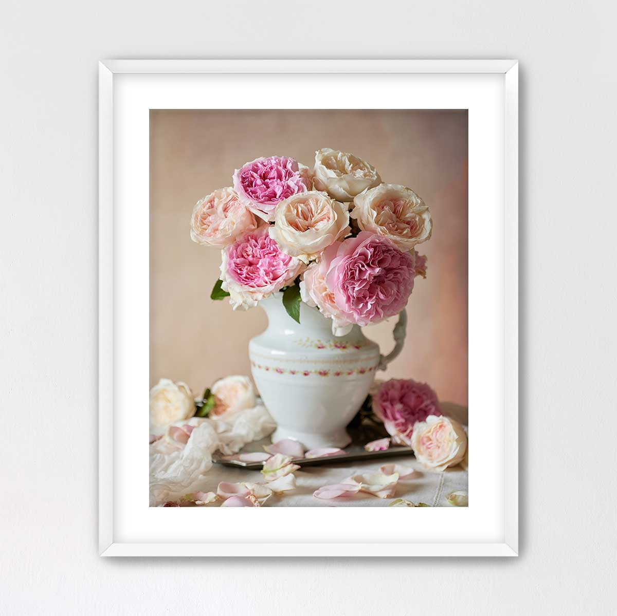 A white vase filled with pink and white roses sits on a table, surrounded by additional rose petals.