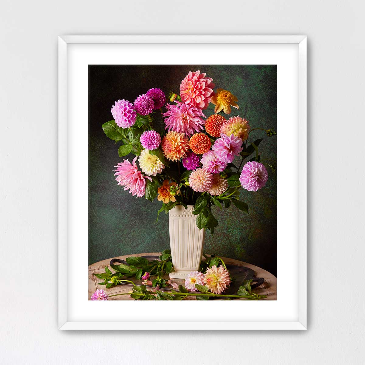 A white vase filled with a vibrant bouquet of pink, orange, and yellow flowers sits on a wooden surface against a dark green background.