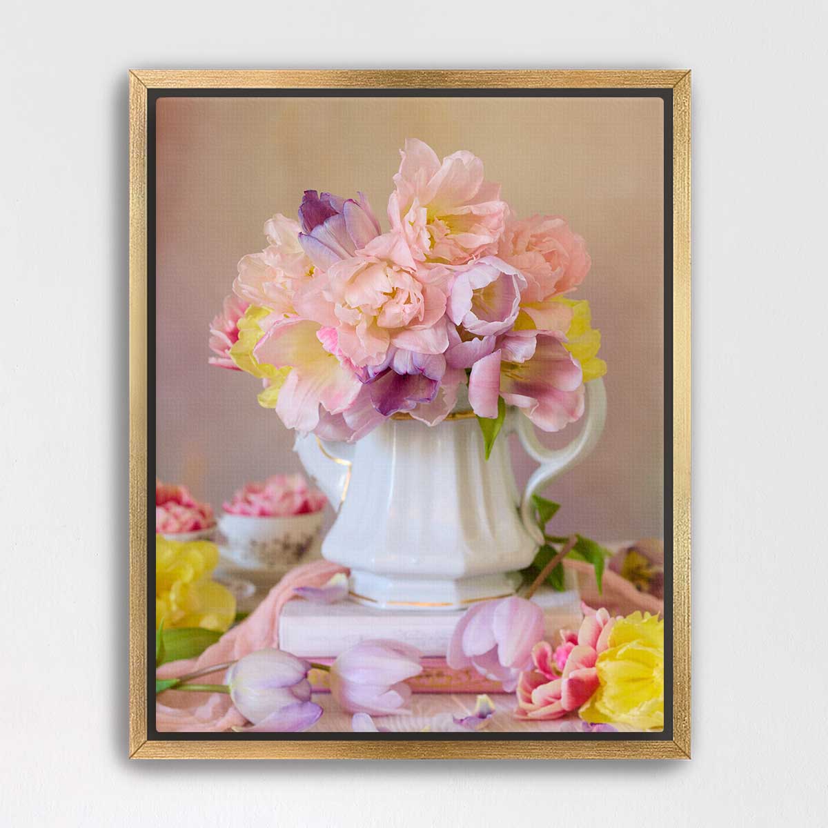 A white vase filled with pink and yellow flowers sits on a table, surrounded by additional flowers and a book.